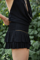 Woman wearing TIC TOC SL31886 VIERRA gauze smocked stud ruffle mini skirt featuring lightweight crinkle gauze fabric, a tiered ruffle mini silhouette, gold stud trim along the waist, smocked elastic waistband at the back, and built-in shorts for coverage, styled for a western-inspired summer festival look.
