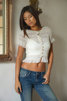 Woman wearing TIC TOC TL31686 RUELLE cropped short sleeve shirred chiffon top featuring allover shirred stretch texture, a soft boat neckline, delicate ruffle trim on the sleeves, subtle ruffle hem detail, and faux pearly button accents for a feminine summer look.