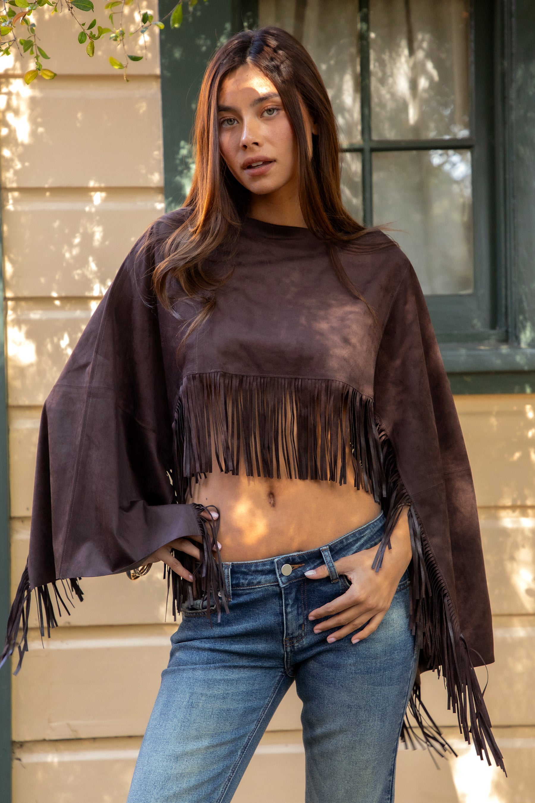 Woman wearing TIC TOC TL31825 STALLION faux suede fringe poncho featuring a cropped cape silhouette, drop shoulder relaxed fit, long fringe tassel detailing along the hem and sleeves, and a soft western-inspired faux suede texture, styled over a fitted top with denim bottoms for a rodeo festival look.