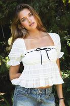 Woman wearing WINSLET square neck pleated ruched chiffon short sleeve top featuring lightweight pleated chiffon fabric, lace trim square neckline, subtle bow detail at the bust, soft ruffle sleeves, ruched texture, and a smocked back panel with adjustable tie for a flexible fit.