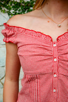 Woman wearing MAREN gingham off shoulder ruffle cap sleeve top featuring a soft ruffle neckline, ruched bust detail, functional button front, smocked back panel, ruffled peplum hem, and lightweight cotton gingham fabric for feminine summer styling.