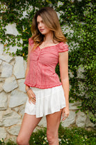Woman wearing MAREN gingham off shoulder ruffle cap sleeve top featuring a soft ruffle neckline, ruched bust detail, functional button front, smocked back panel, ruffled peplum hem, and lightweight cotton gingham fabric for feminine summer styling.