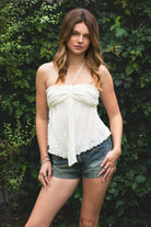 Woman wearing ODETTE lace handkerchief halter top featuring soft textured lace fabric, twisted bandeau bodice, adjustable halter neck tie, romantic handkerchief hemline, delicate lace trim, ruched texture, and lightweight lining for summer vacation and festival styling.