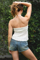 Woman wearing ODETTE lace handkerchief halter top featuring soft textured lace fabric, twisted bandeau bodice, adjustable halter neck tie, romantic handkerchief hemline, delicate lace trim, ruched texture, and lightweight lining for summer vacation and festival styling.
