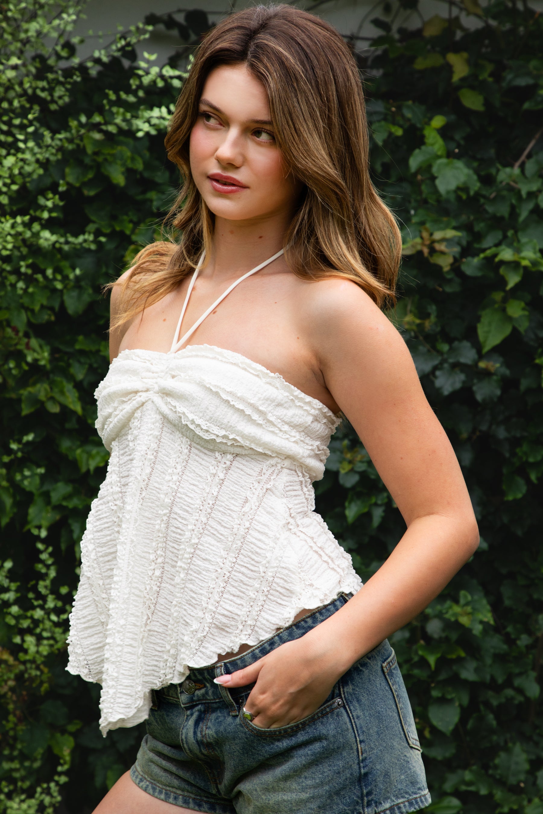 Woman wearing ODETTE lace handkerchief halter top featuring soft textured lace fabric, twisted bandeau bodice, adjustable halter neck tie, romantic handkerchief hemline, delicate lace trim, ruched texture, and lightweight lining for summer vacation and festival styling.
