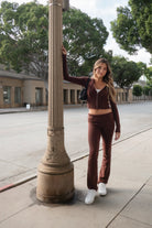 Woman wearing TIC TOC TPC29297-COZEE brown sweater knit two-piece set featuring a cropped zip-up hoodie with ribbed details and matching fold-over flare pants, styled outdoors with sneakers and a small black shoulder bag.