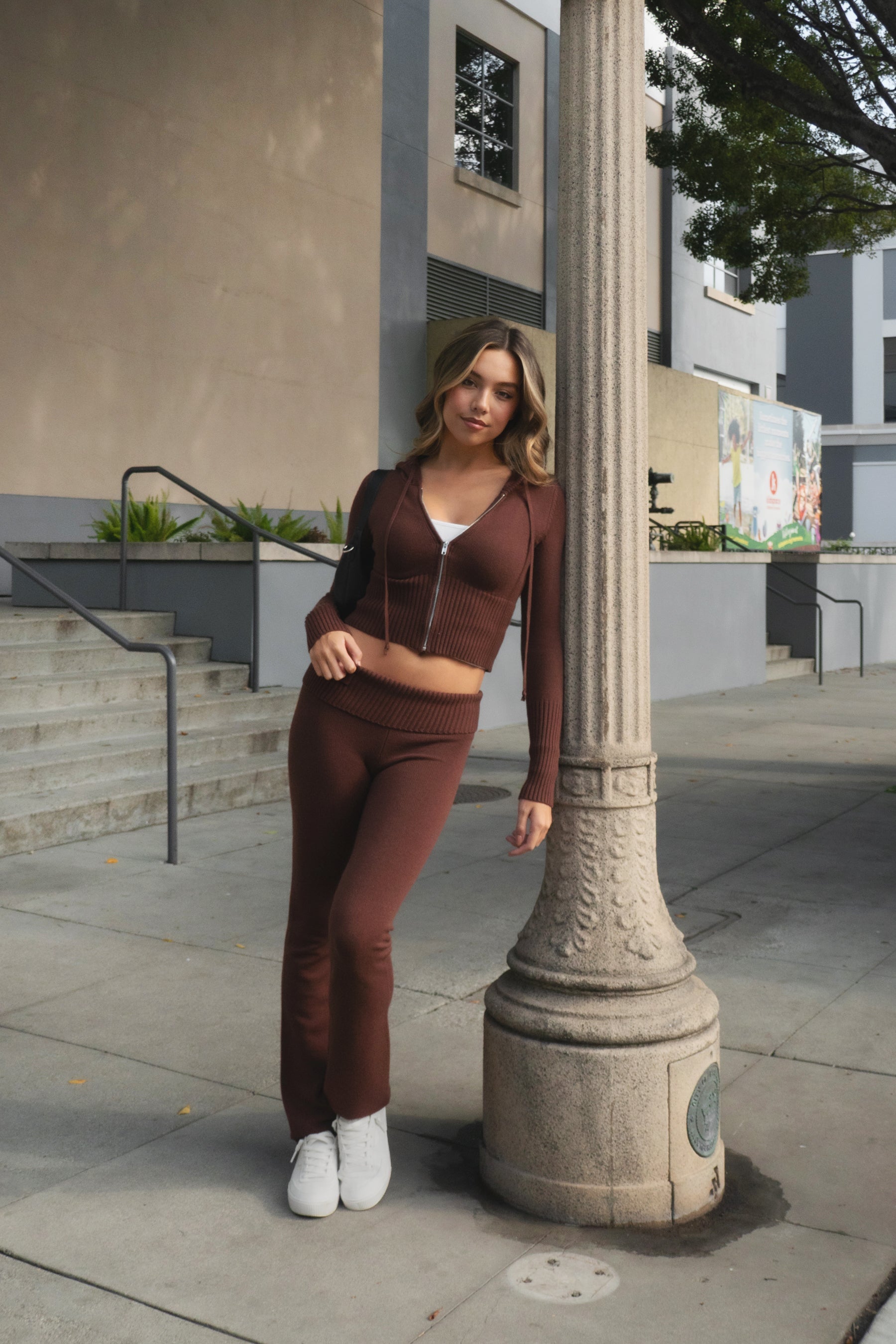 Woman wearing TIC TOC TPC29297-COZEE brown sweater knit two-piece set featuring a cropped zip-up hoodie with ribbed details and matching fold-over flare pants, styled outdoors with sneakers and a small black shoulder bag.