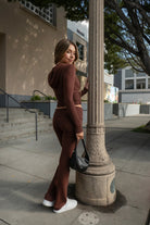 Woman wearing TIC TOC TPC29297-COZEE brown sweater knit two-piece set featuring a cropped zip-up hoodie with ribbed details and matching fold-over flare pants, styled outdoors with sneakers and a small black shoulder bag.