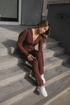 Woman wearing TIC TOC TPC29297-COZEE brown sweater knit two-piece set featuring a cropped zip-up hoodie with ribbed details and matching fold-over flare pants, styled outdoors with sneakers and a small black shoulder bag.