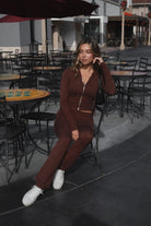 Woman wearing TIC TOC TPC29297-COZEE brown sweater knit two-piece set featuring a cropped zip-up hoodie with ribbed details and matching fold-over flare pants, styled outdoors with sneakers and a small black shoulder bag.