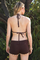 Woman wearing CORALYN crochet seashell bikini top and short set featuring a halter triangle top with self tie neck and back, seashell charm trim along the underbust, mid-rise crochet shorts with elastic waistband, and seashell and bead trim at the hem in a soft textured cotton knit.