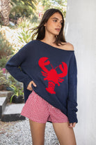 Woman wearing LOBELLE lobster graphic off shoulder sweater featuring relaxed sweater knit fabric, bold contrast red lobster intarsia graphic at the front, wide neckline designed to drape off the shoulder, oversized slouchy silhouette, dropped shoulders, long relaxed sleeves, ribbed cuffs and hem, and subtle side slits.