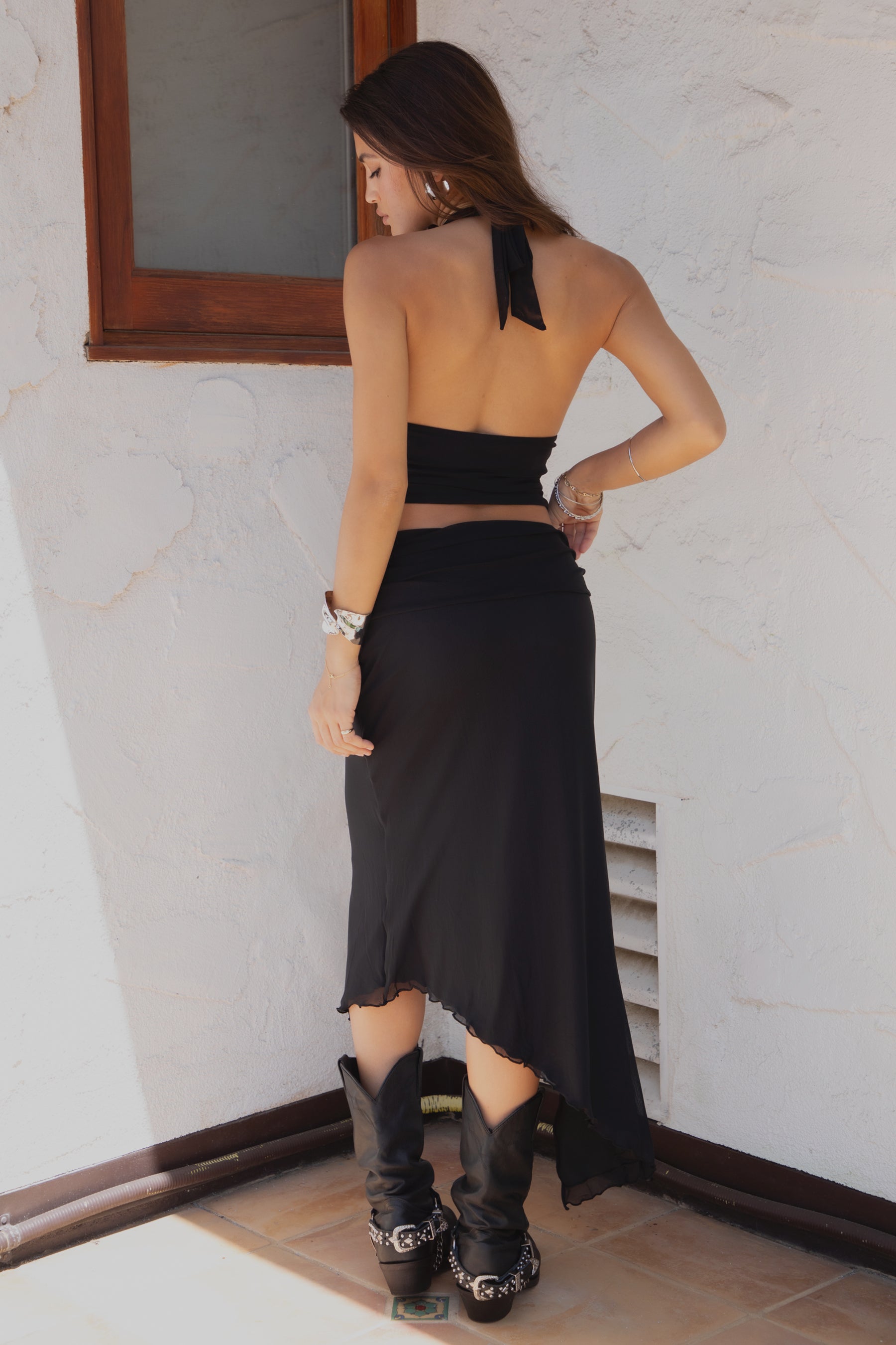 Woman wearing TIC TOC ALMEDA Mesh Print Halter and Maxi Skirt Set in black with studded detail, scarf hem top, and high slit skirt paired with black cowboy boots against a white wall.
