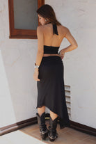 Woman wearing TIC TOC ALMEDA Mesh Print Halter and Maxi Skirt Set in black with studded detail, scarf hem top, and high slit skirt paired with black cowboy boots against a white wall.