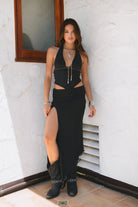 Woman wearing TIC TOC ALMEDA Mesh Print Halter and Maxi Skirt Set in black with studded detail, scarf hem top, and high slit skirt paired with black cowboy boots against a white wall.