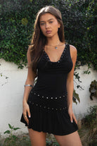 Woman wearing TIC TOC TSH32013-RAVELLE RAVELLE two piece skirt set featuring a cowl neck sleeveless top with soft draped front and eyelet trim along the neckline and waist, paired with a high waist mini flare skirt with studded western accents and soft movement in smooth stretch jersey, styled with boots for a festival-ready western summer look.