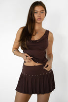 Woman wearing TIC TOC TSH32013-RAVELLE RAVELLE two piece skirt set featuring a cowl neck sleeveless top with soft draped front and eyelet trim along the neckline and waist, paired with a high waist mini flare skirt with studded western accents and soft movement in smooth stretch jersey, styled with boots for a festival-ready western summer look.