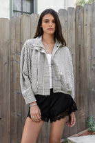 Woman wearing MARGOT plaid gingham zip up bomber jacket featuring lightweight crinkled cotton blend fabric, classic gingham plaid pattern, high neck collar, front button and zip closure, semi cropped length, sleeve snap buttons, relaxed drop shoulder silhouette, side pockets, and elastic hem.
