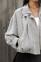 Woman wearing MARGOT plaid gingham zip up bomber jacket featuring lightweight crinkled cotton blend fabric, classic gingham plaid pattern, high neck collar, front button and zip closure, semi cropped length, sleeve snap buttons, relaxed drop shoulder silhouette, side pockets, and elastic hem.