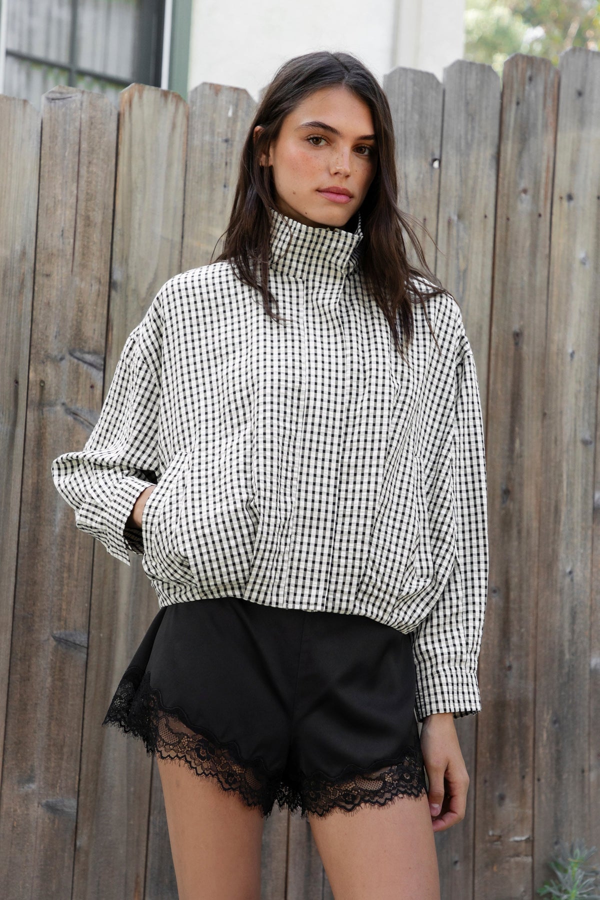 Woman wearing MARGOT plaid gingham zip up bomber jacket featuring lightweight crinkled cotton blend fabric, classic gingham plaid pattern, high neck collar, front button and zip closure, semi cropped length, sleeve snap buttons, relaxed drop shoulder silhouette, side pockets, and elastic hem.