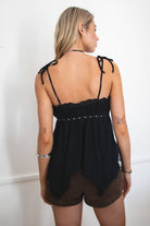Woman wearing TIC TOC GLIMSHA black mesh boho cami top featuring adjustable tie straps, frill neckline, studded trim, and handkerchief hem with brown mini shorts