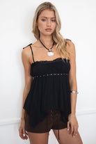 Woman wearing TIC TOC GLIMSHA black mesh boho cami top featuring adjustable tie straps, frill neckline, studded trim, and handkerchief hem with brown mini shorts