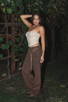 Woman wearing the TIC TOC PF31461 IZLA brown denim twill mid rise studded carpenter pants featuring double-knee seams, wide-leg fit, and vintage faded wash paired with an embroidered cream strapless top in an outdoor setting. 