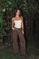 Woman wearing the TIC TOC PF31461 IZLA brown denim twill mid rise studded carpenter pants featuring double-knee seams, wide-leg fit, and vintage faded wash paired with an embroidered cream strapless top in an outdoor setting. 