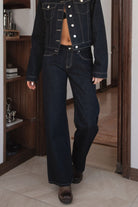 Woman wearing TIC TOC PF31703 MILLS selvedge denim straight leg pants in dark blue wash with a low-rise waist and wide-leg silhouette with a matching jackte in an indoor setting. 