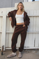 Person wearing a brown tracksuit and white crop top in front of a white wooden door.