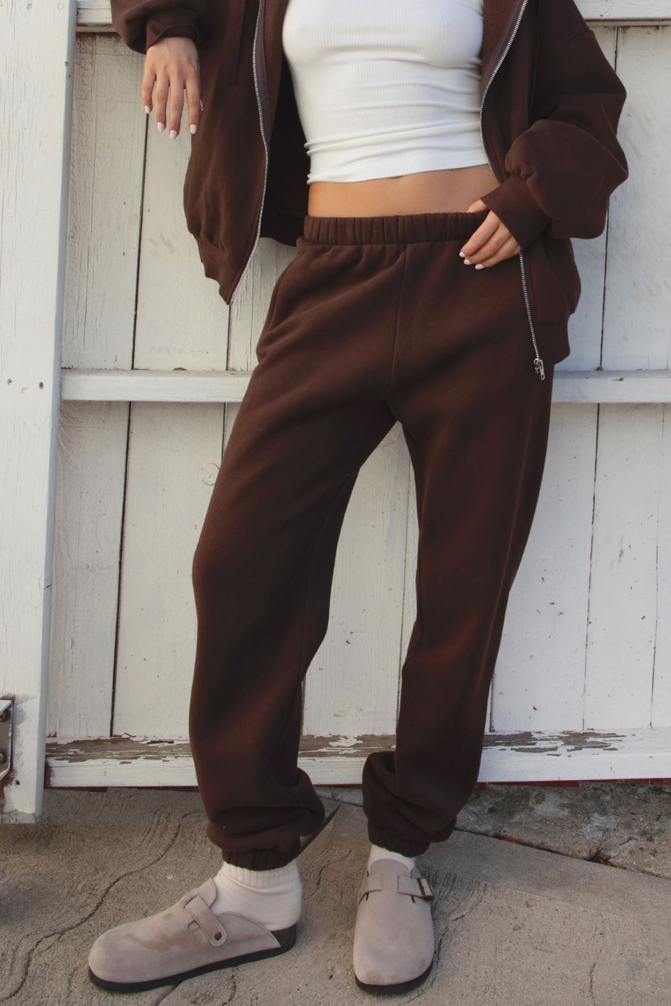 Person wearing a brown tracksuit and white crop top in front of a white wooden door.