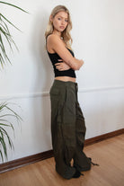 Woman wearing a black tank top and TIC TOC PH29131 TOMY olive crinkled nylon parachute cargo pants with adjustable drawstring waistband and wide-leg silhouette against a white wall.