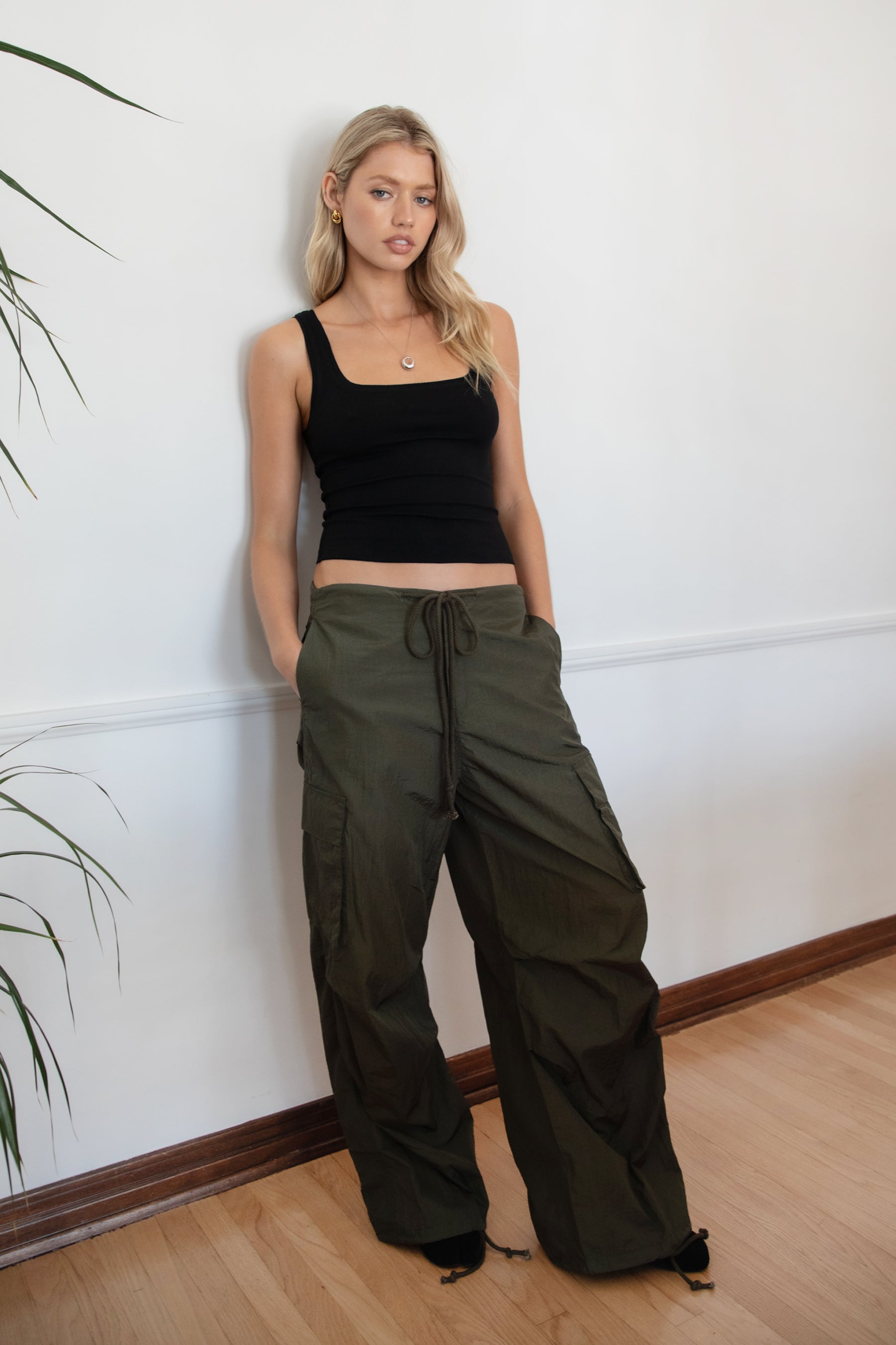 Woman wearing a black tank top and TIC TOC PH29131 TOMY olive crinkled nylon parachute cargo pants with adjustable drawstring waistband and wide-leg silhouette against a white wall.