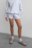 Woman wearing the TIC TOC PJ31004 CARLY ash cloud fleece low rise shorts featuring an elastic waistband with drawcord, deep side pockets, and raw cut hemline paired with a matching top