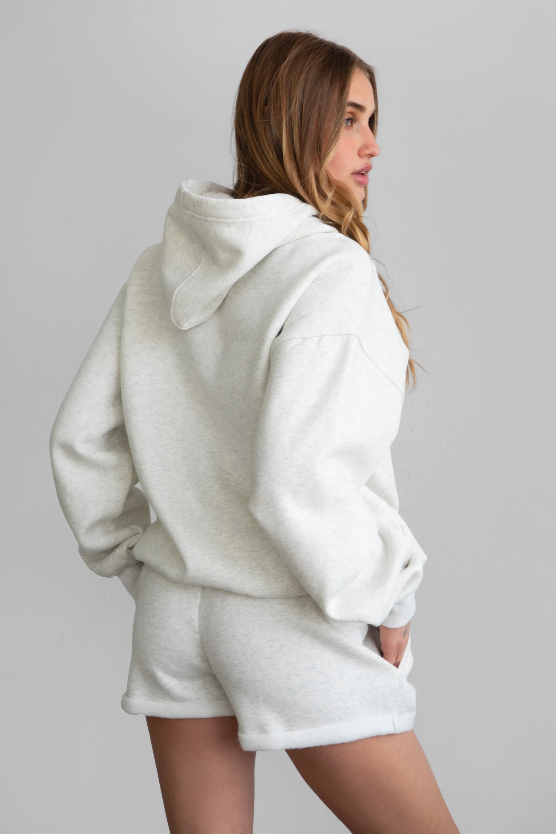 Woman wearing the TIC TOC PJ31004 CARLY heather white cloud fleece low rise shorts featuring an elastic waistband with drawcord, deep side pockets, and raw cut hemline paired with a matching top