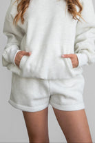 Woman wearing the TIC TOC PJ31004 CARLY heather white cloud fleece low rise shorts featuring an elastic waistband with drawcord, deep side pockets, and raw cut hemline paired with a matching top