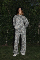 Woman wearing TIC TOC PJ31369 ELK woodland camo fleece wide leg sweatpants with elastic waistband, drawcord, and side pockets with matching top standing in grass with a bush in the background