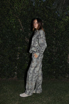 Woman wearing TIC TOC PJ31369 ELK woodland camo fleece wide leg sweatpants with elastic waistband, drawcord, and side pockets with matching top standing in grass with a bush in the background