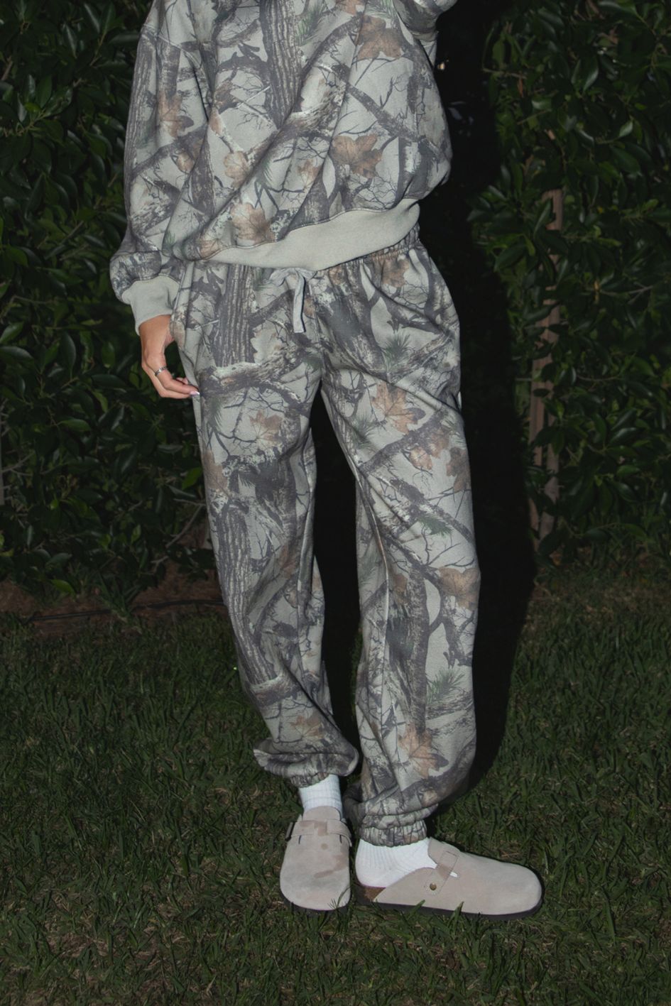 Woman wearing TIC TOC PJ31425 ELK woodland camo fleece jogger sweatpants with elastic tie waistband and tapered cinched ankles with matching top standing against a dark background