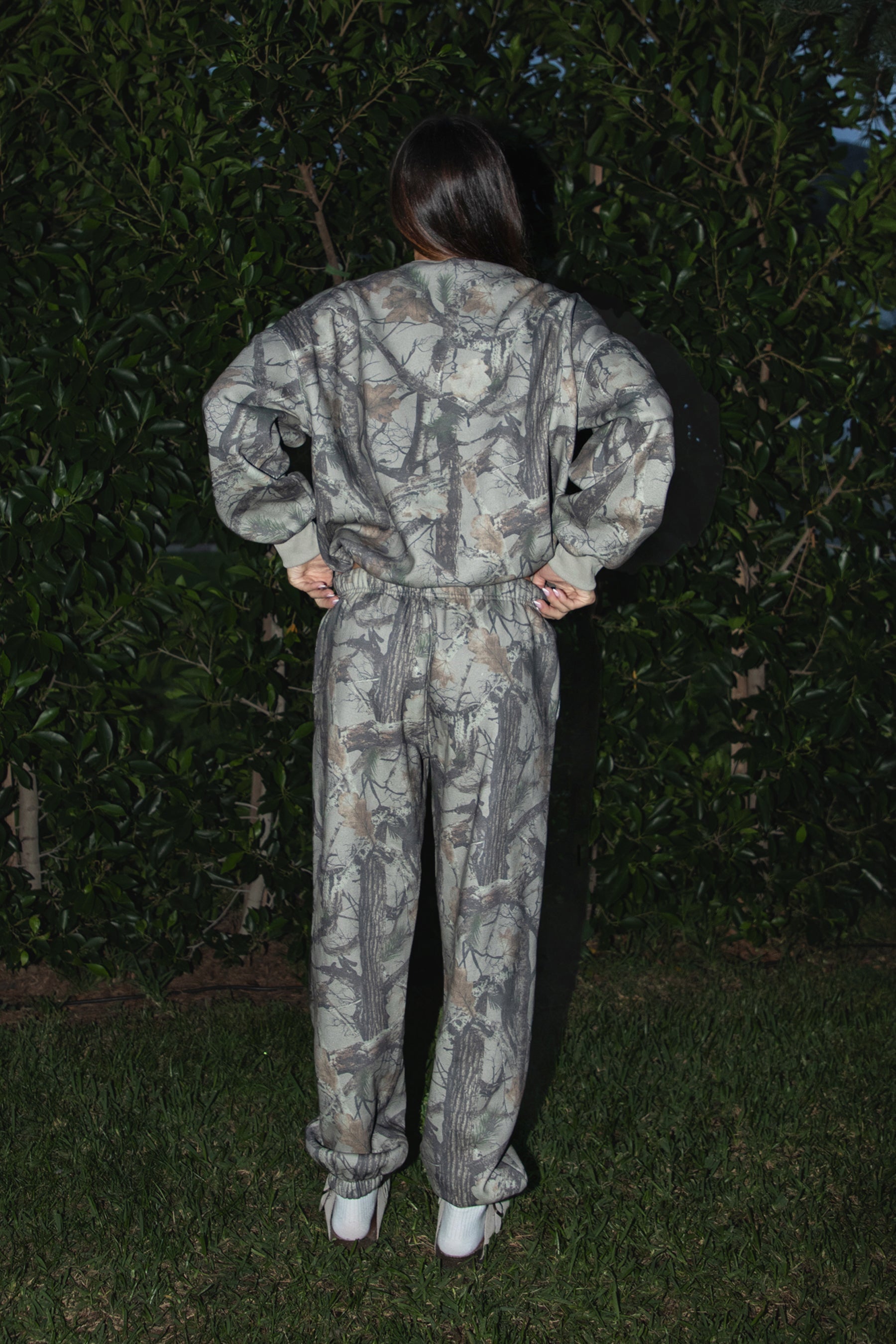 Woman wearing TIC TOC PJ31425 ELK woodland camo fleece jogger sweatpants with elastic tie waistband and tapered cinched ankles with matching top standing against a dark background