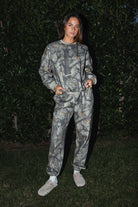 Woman wearing TIC TOC PJ31425 ELK woodland camo fleece jogger sweatpants with elastic tie waistband and tapered cinched ankles with matching top standing against a dark background