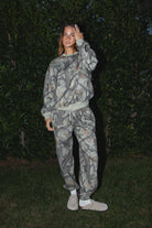 Woman wearing TIC TOC PJ31425 ELK woodland camo fleece jogger sweatpants with elastic tie waistband and tapered cinched ankles with matching top standing against a dark background
