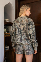Woman wearing a camouflage outfit standing in a room with wooden door and shelves.
