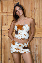 Woman wearing the TIC TOC SA31601 MOOEDY faux fur cow print mini skirt featuring whipstitch suede trim, side zipper, and built-in shorts. with matching top against a wooden fence background