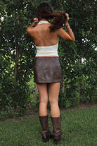 Woman wearing the TIC TOC SH31455 DAYNA brown PU leather low rise mini skirt featuring faux side zippers, button closure, and functional pockets in stretch leather fabric paired with a cream cowl neck top and knee high boots. 