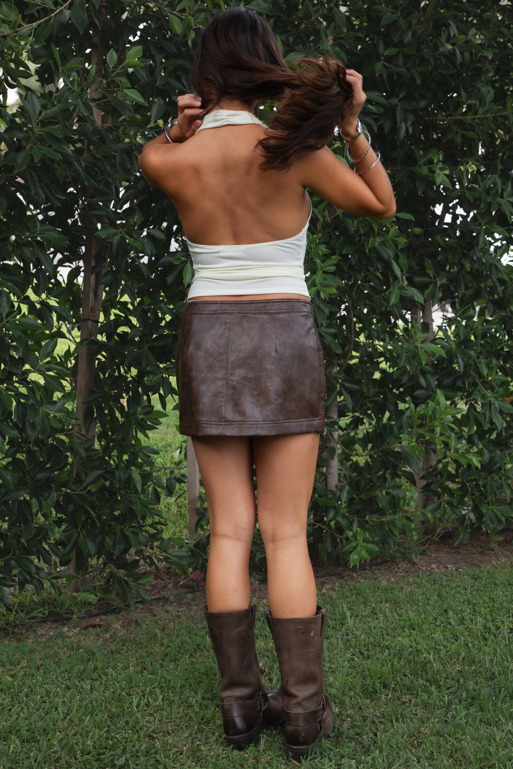 Woman wearing the TIC TOC SH31455 DAYNA brown PU leather low rise mini skirt featuring faux side zippers, button closure, and functional pockets in stretch leather fabric paired with a cream cowl neck top and knee high boots. 