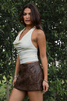 Woman wearing the TIC TOC SH31455 DAYNA brown PU leather low rise mini skirt featuring faux side zippers, button closure, and functional pockets in stretch leather fabric paired with a cream cowl neck top.
