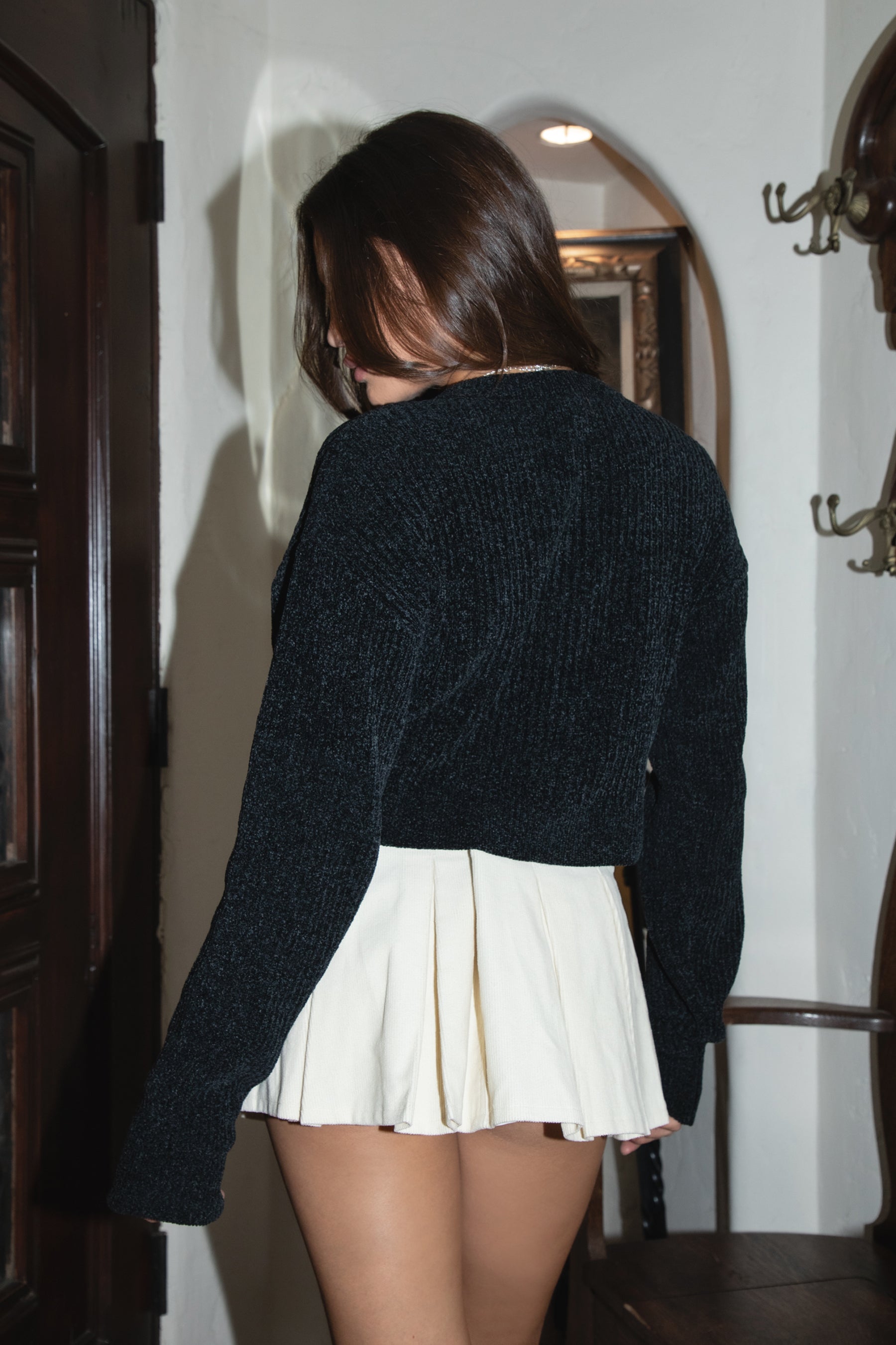 Person wearing a black sweater and white pleated skirt in a room with wooden furniture.