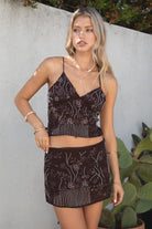 Model wearing the TIC TOC SL31480 VEDA brown chiffon beaded low-rise mini skort featuring hand-beaded floral embroidery, chiffon overlay, and built-in shorts with matching cami top in an outdoor setting. 