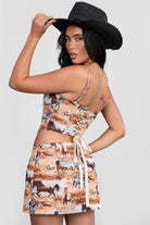A model wearing a brown western-themed horse print crop top with adjustable shoulder straps and a square neckline. The top is paired with a matching skirt and accessorized with a wide-brimmed hat and a necklace.